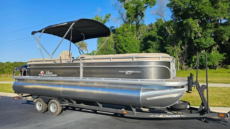 The Image of 2024 Sun Tracker Party Barge 24 DLX pontoon boat on a trailer, parked outdoors. - 0