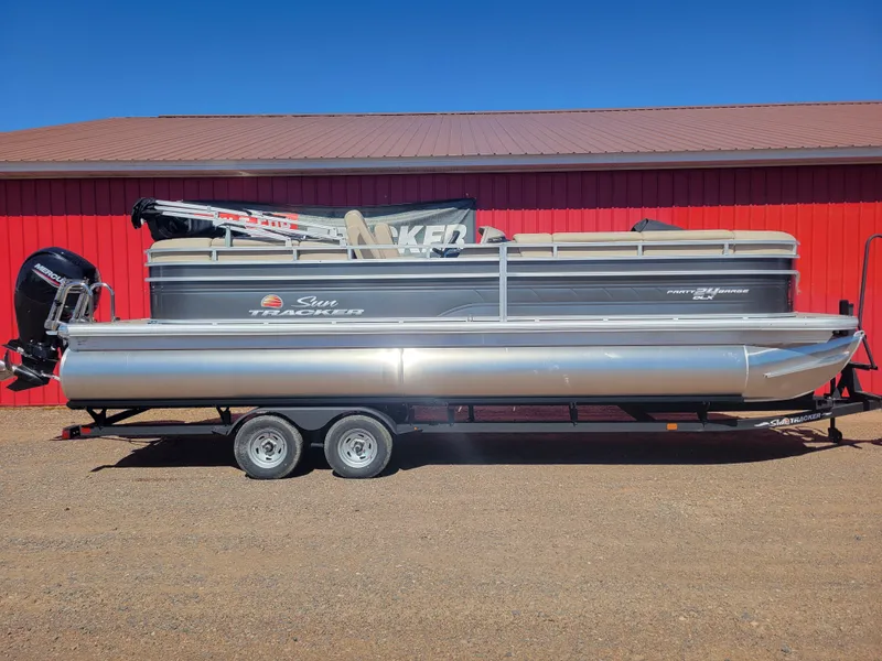 The Image of 2024 Sun Tracker Party Barge 24 DLX pontoon boat on trailer, red building background. - 0