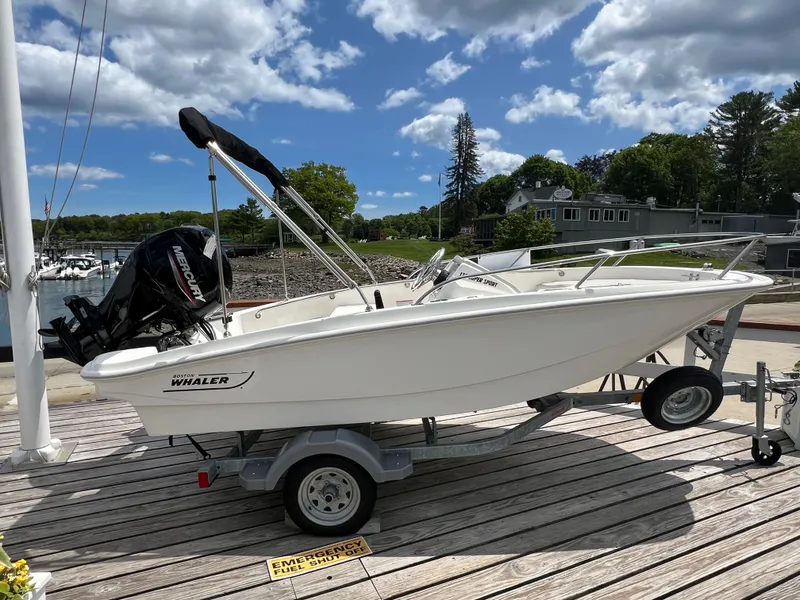 Slide: The Image of 2024 Boston Whaler 130 Super Sport boat on trailer, docked under a partly cloudy sky. - 0