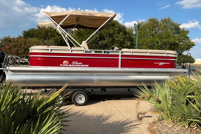 Slide: The Image of 2024 Sun Tracker Party Barge 24 DLX pontoon boat on trailer, surrounded by greenery. - 2