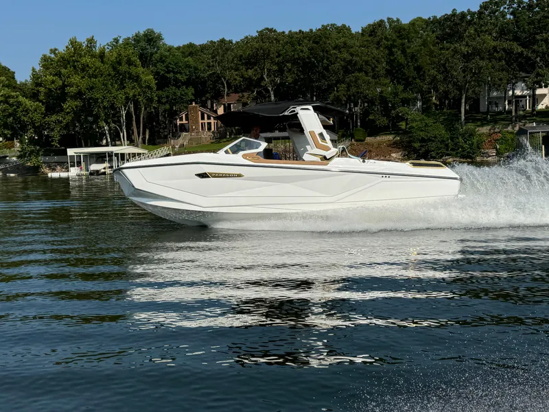 The Image of 2024 Nautique G25 Paragon boat cruising on a lake with lush green shoreline. - 0