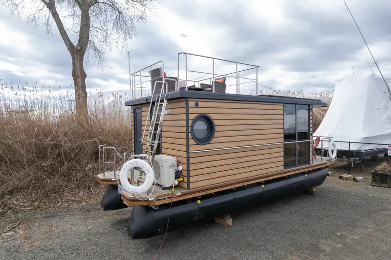 Slide: The Image of 2024 Waterlodge Apart M houseboat with wooden exterior and rooftop deck, docked on a gravel shore. - 13