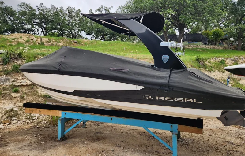 Slide: The Image of 2016 Regal 2100 Bowrider boat covered and on a stand, surrounded by greenery. - 2