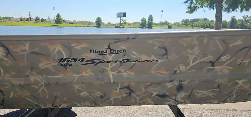 Slide: The Image of 2024 Tracker Grizzly 1654 T Sportsman boat with camouflage design by a lake. - 2