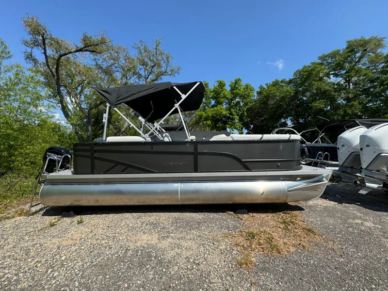 Slide: The Image of 2024 Crest Classic LX 220 pontoon boat with black canopy on gravel lot. - 1