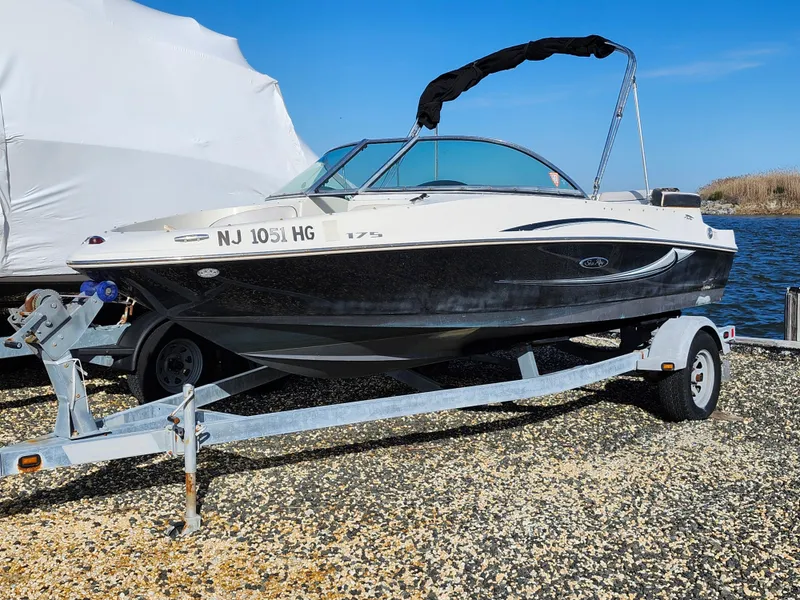 The Image of 2010 Sea Ray 175 Sport boat on trailer, parked on gravel near water. - 0