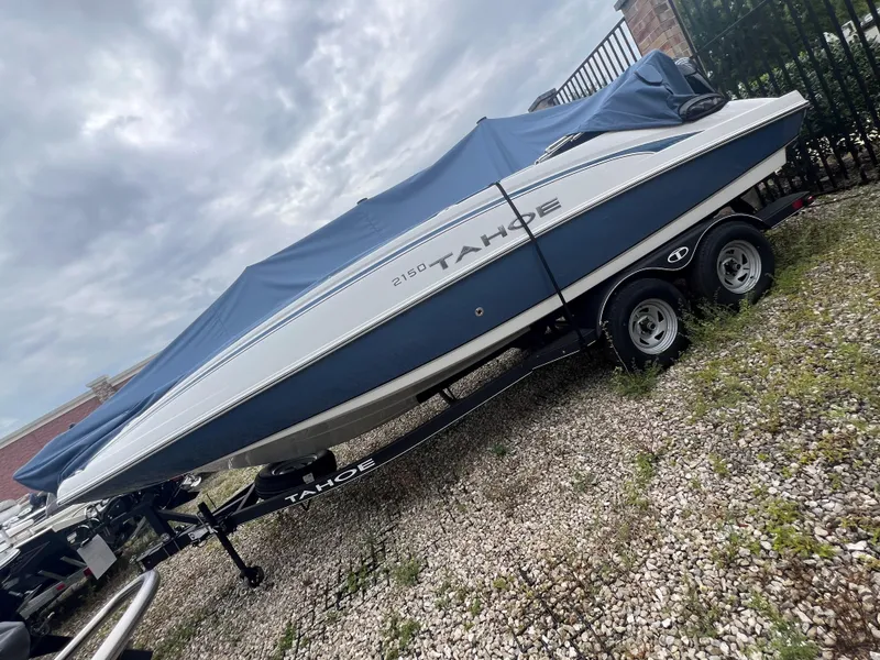 Slide: The Image of 2024 Tahoe 2150 boat with blue cover on trailer, parked on gravel. - 0