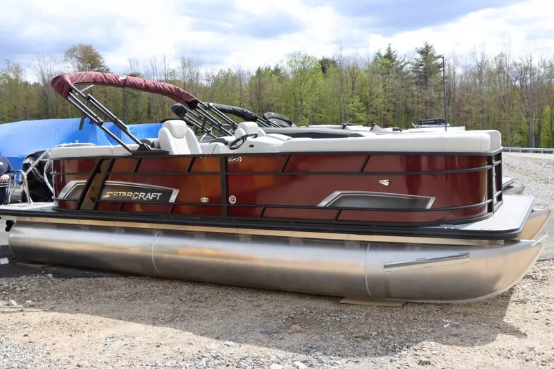 Slide: The Image of 2024 Starcraft EXS 3 pontoon boat with maroon exterior, parked on gravel. - 1