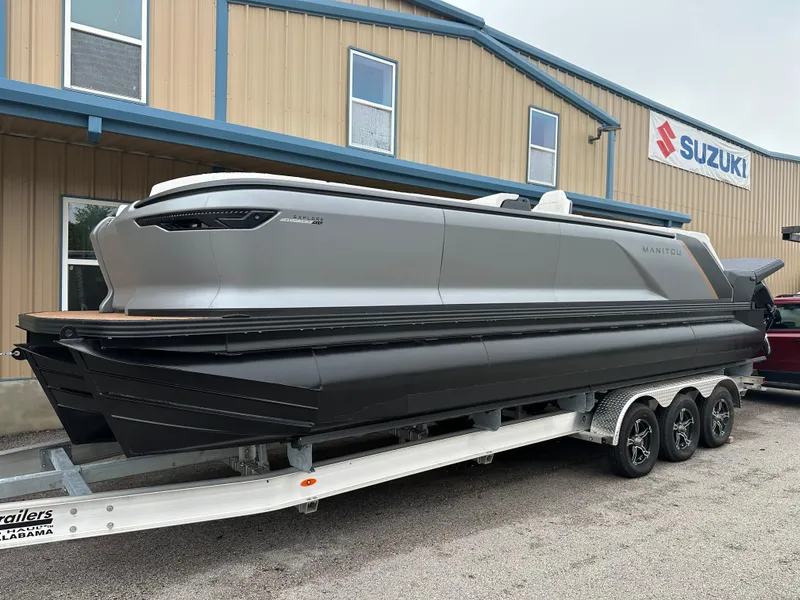 The Image of 2024 Manitou Explore Navigator Dual Engine Rotax pontoon boat on a trailer outside a Suzuki dealership. - 0