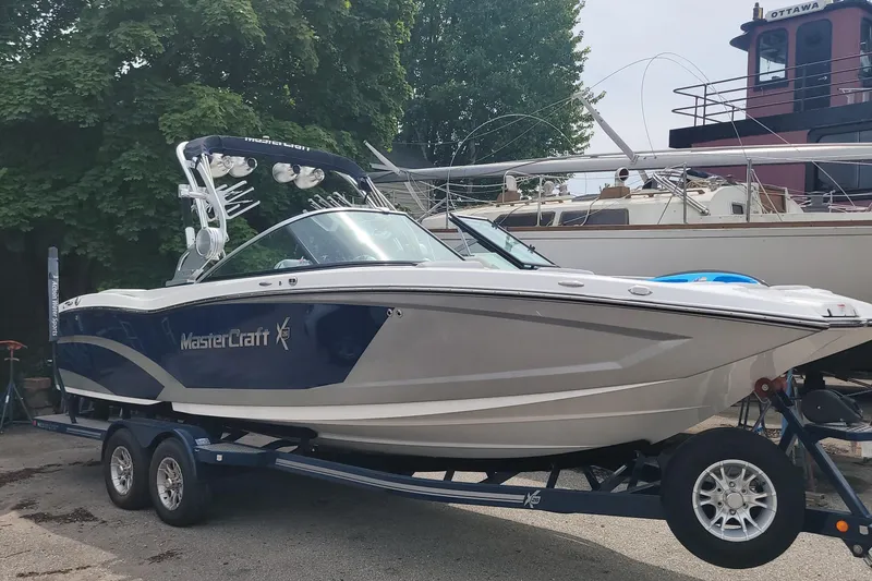 The Image of 2016 MasterCraft X26 boat on a trailer, parked near other boats and trees. - 0