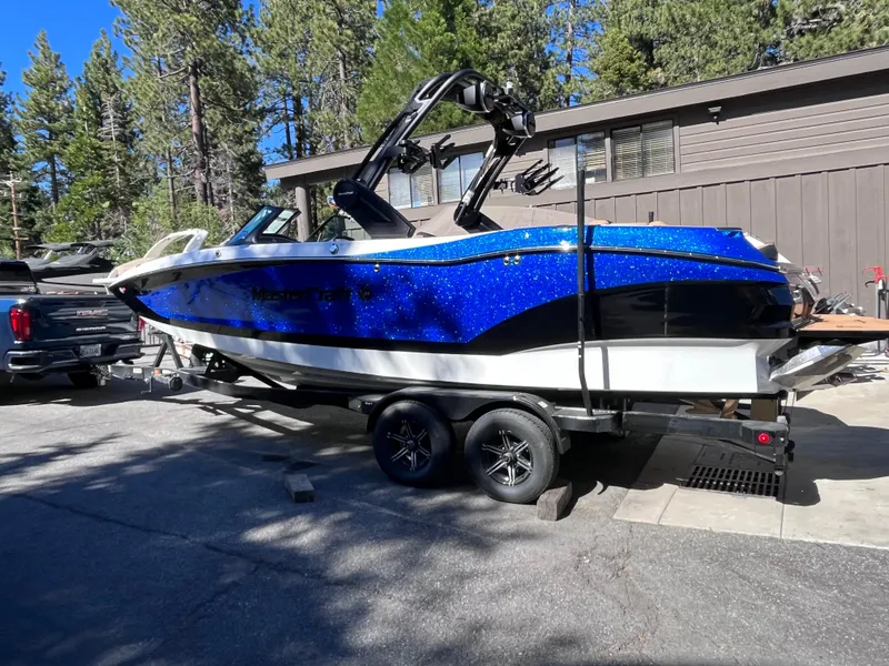 Slide: The Image of 2024 MasterCraft X24 boat on trailer, blue and white, parked outdoors near trees and building. - 3