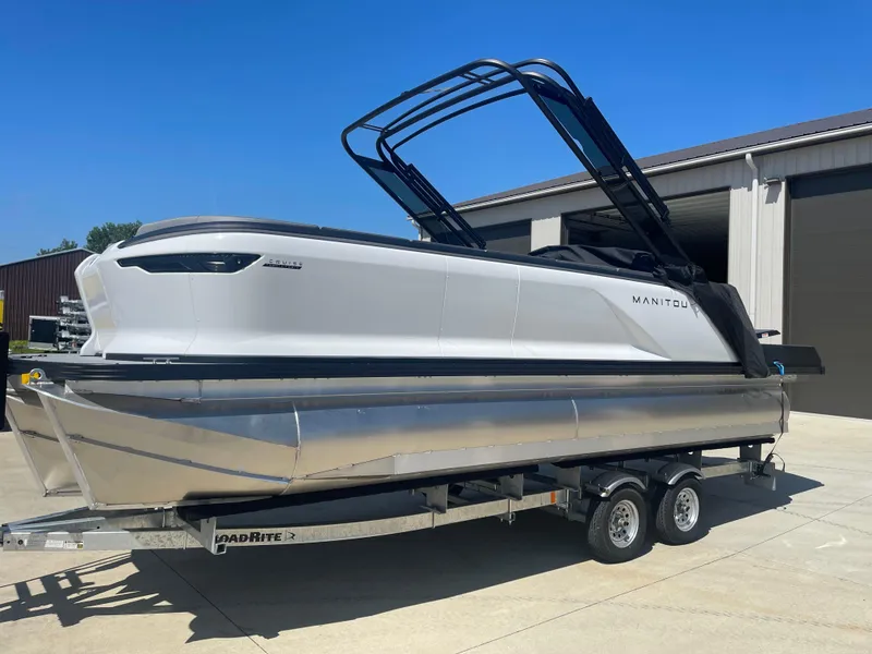 Slide: The Image of 2024 Manitou Cruise pontoon boat on a trailer outside a warehouse. - 2
