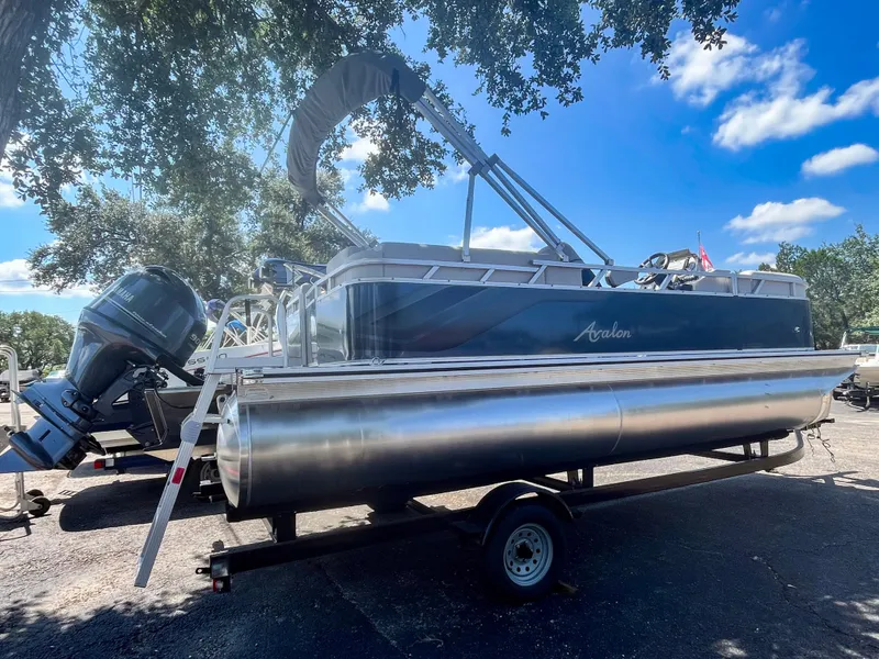 Slide: The Image of 2024 Avalon VLS 21 Quad Lounger pontoon boat on trailer under trees. - 2