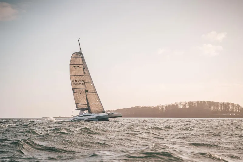 Slide: The Image of Sailing boat Dragonfly 28, 2026 model, gliding on open water near a wooded shoreline. - 0