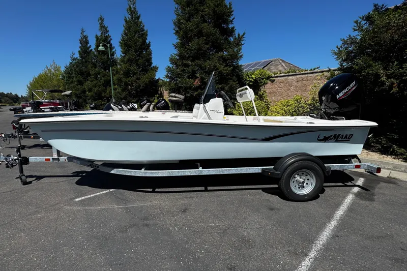 The Image of A 2024 Mako Pro Skiff 17 CC on a trailer in a parking lot, surrounded by trees. - 0