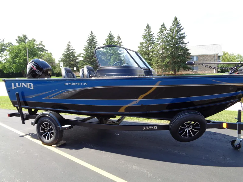 Slide: The Image of 2024 Lund 1875 Impact XS boat on trailer, black and blue, parked outdoors. - 18