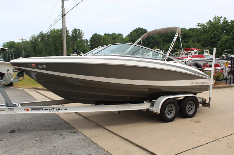 Slide: The Image of 2011 Regal 2100 boat on trailer, parked outdoors, surrounded by trees and other boats. - 2