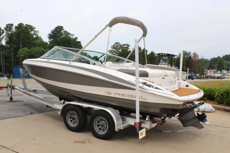 Slide: The Image of 2011 Regal 2100 boat on trailer, parked outdoors, surrounded by trees and marina. - 3