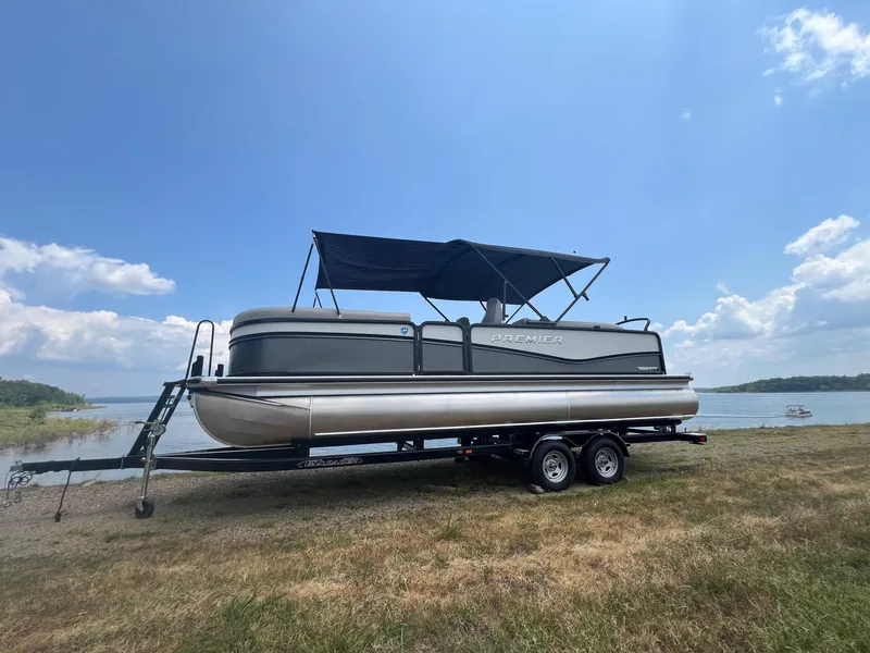 Slide: The Image of 2024 Premier 230 Sunsation RE pontoon boat on trailer by a lake. - 0