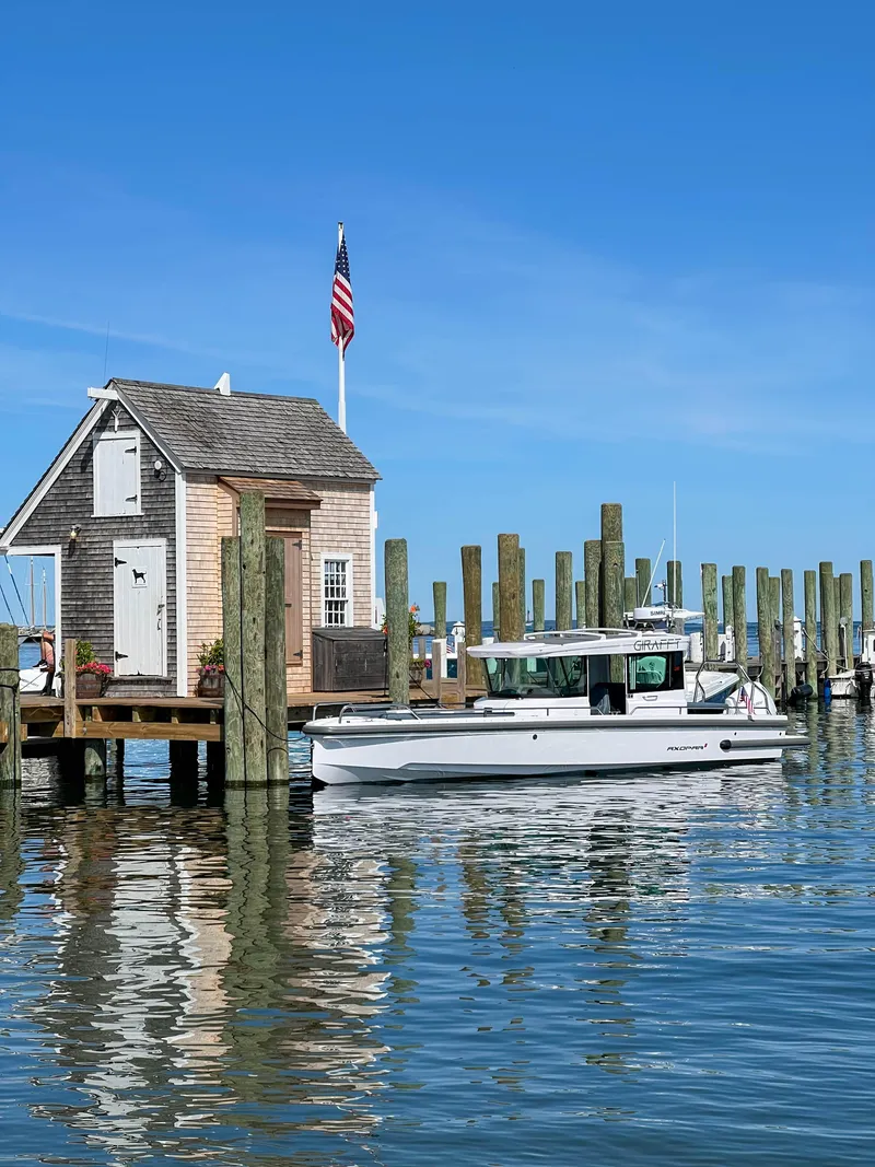 Slide: The Image of 2022 Axopar 28 Cabin Aft Cabin boat docked near a quaint wooden boathouse. - 2