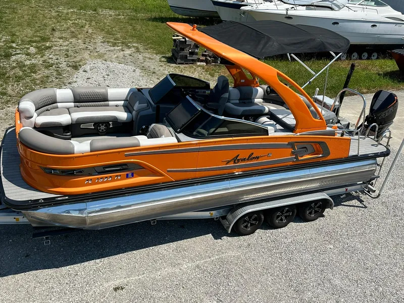 Slide: The Image of 2023 Avalon Excalibur Elite Windshield pontoon boat on a trailer, orange and silver design. - 2