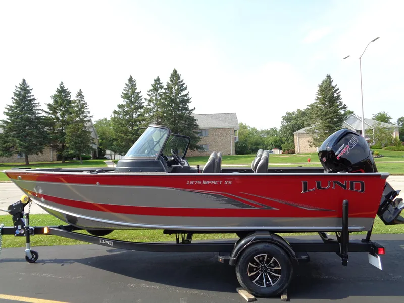 The Image of 2024 Lund 1875 Impact XS boat, red and silver, on trailer, parked outdoors. - 0