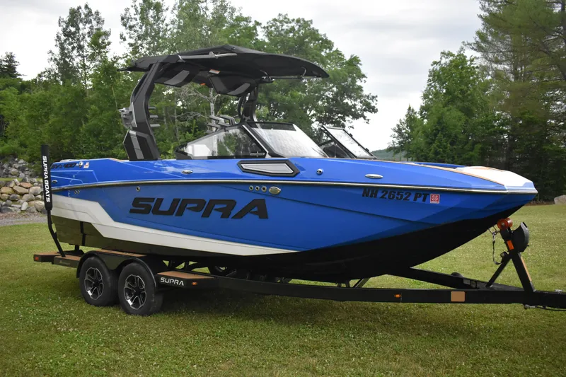 Slide: The Image of 2024 Supra SV boat in blue on a trailer, parked on grass with trees in the background. - 2