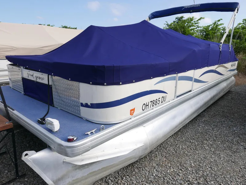 Slide: The Image of 2006 Sweetwater SW2222DF pontoon boat with blue cover, docked on gravel. - 2