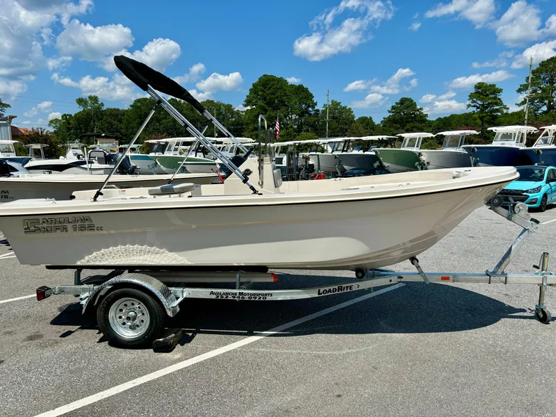 Slide: The Image of 2025 Carolina Skiff 162 JLS boat on trailer, parked outdoors under a clear blue sky. - 6