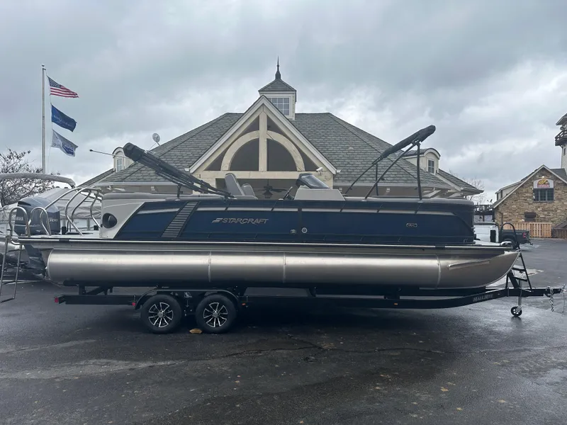 Slide: The Image of 2024 Starcraft MX 25 R pontoon boat on a trailer, parked near a building. - 0