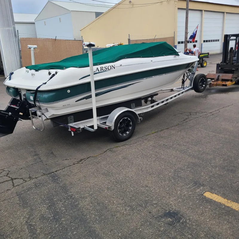 Slide: The Image of 1999 Larson 206 boat on trailer with green cover, parked in industrial area. - 2