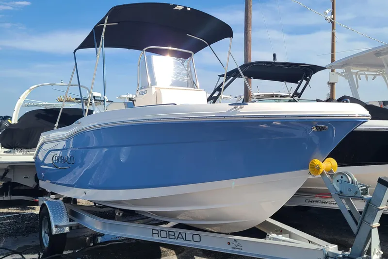 The Image of 2024 Robalo R180 Center Console boat on trailer, blue hull, black canopy. - 0
