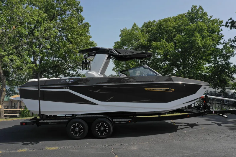 Slide: The Image of 2025 Nautique G23 boat on trailer, parked outdoors with trees in background. - 2