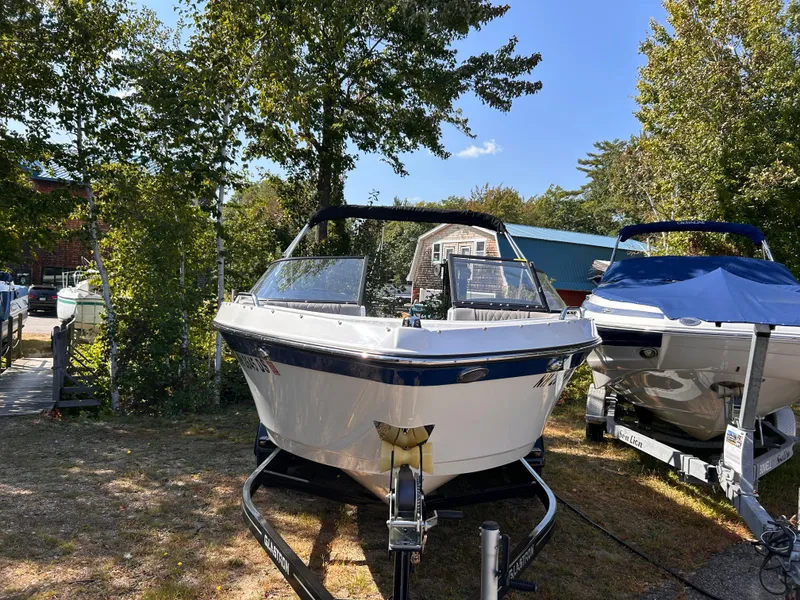 Slide: The Image of 2024 Glastron GX 215 boat on trailer, parked outdoors, surrounded by trees. - 2