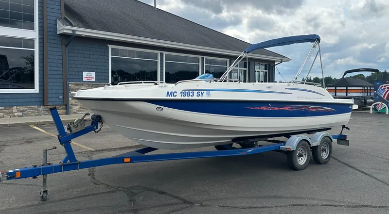 Slide: The Image of 2006 Bayliner 217 boat on trailer, parked outside a building under cloudy skies. - 0