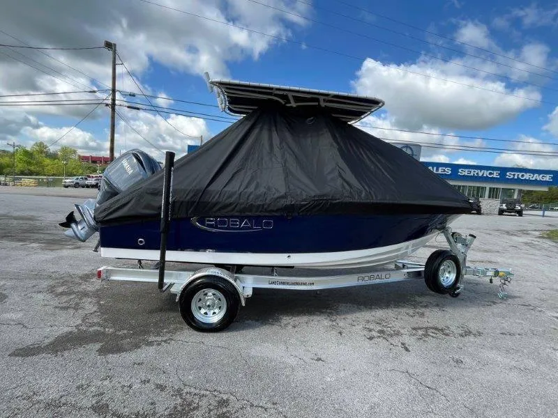 Slide: The Image of 2024 Robalo R180 boat covered on trailer, parked outdoors under cloudy sky. - 13