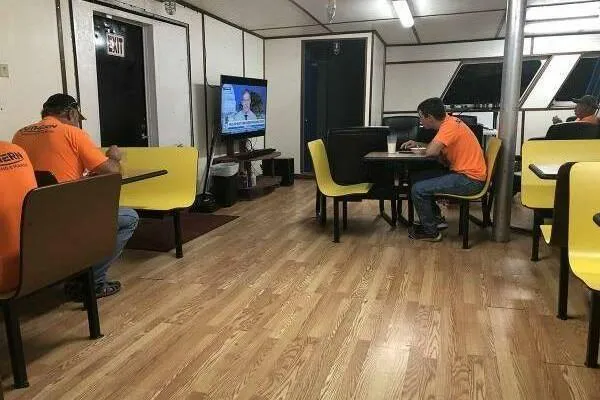 Slide: The Image of Workers in orange shirts relax in a break room with a TV and yellow seating. - 9