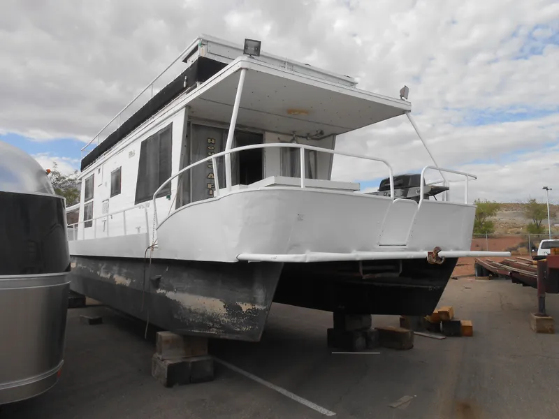 The Image of 1972 Boatel Hurcules houseboat on blocks, white exterior, parked outdoors under cloudy sky. - 1