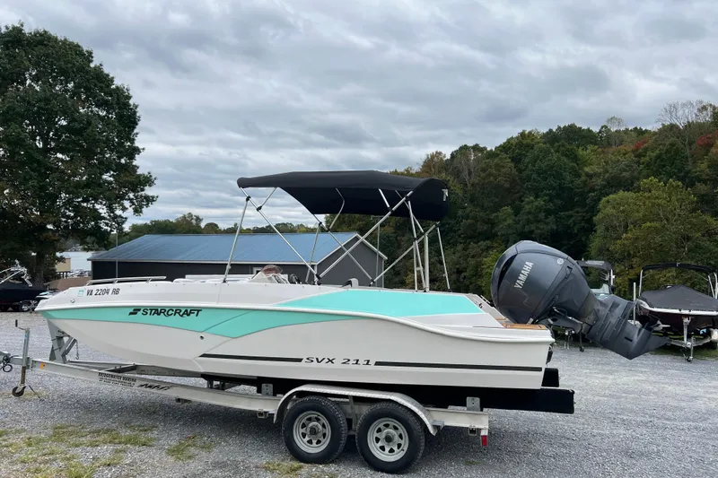 Slide: The Image of 2024 Starcraft SVX 211 OB boat on trailer, parked outdoors with trees in background. - 11
