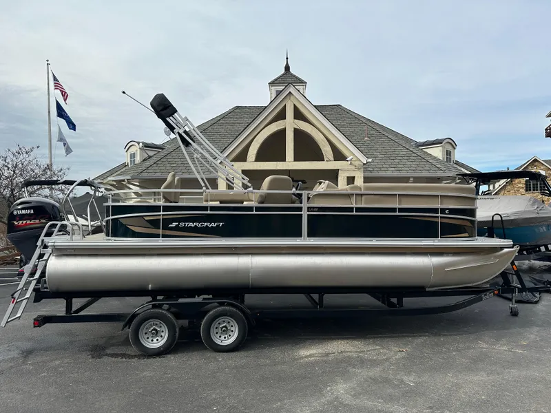 Slide: The Image of 2024 Starcraft LX 22 F pontoon boat on trailer, parked near a building. - 0