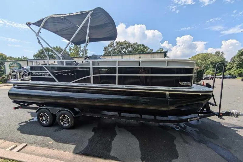 Slide: The Image of 2024 Ranger Reata 223C pontoon boat with canopy on trailer, parked outdoors. - 0