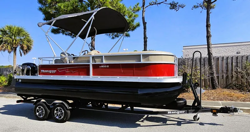 Slide: The Image of 2024 Ranger Reata 200C pontoon boat on trailer, red and black design, parked outdoors. - 2