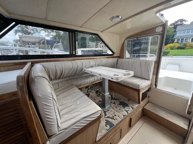 Slide: The Image of 1990 Bertram 28 Flybridge interior with cushioned seating and table, featuring large windows. - 27