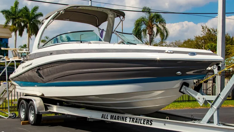 Slide: The Image of 2023 Crownline 290 XSS boat on trailer, parked outdoors with palm trees in background. - 0