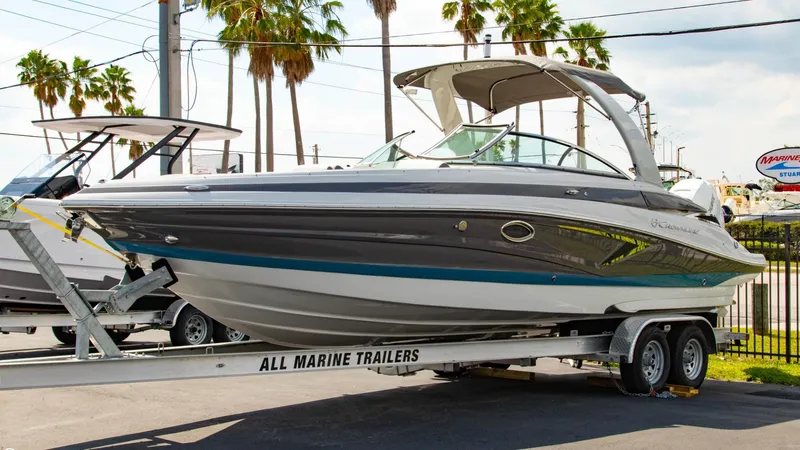 Slide: The Image of 2023 Crownline 290 XSS boat on trailer, parked outdoors with palm trees in background. - 2