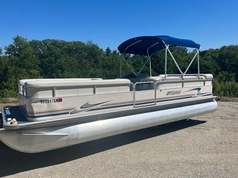 Slide: The Image of 2003 Sylvan Mirage pontoon boat with blue canopy on gravel, surrounded by trees. - 6