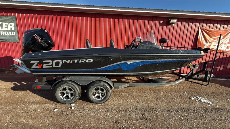 The Image of 2024 Nitro ZV20 boat on trailer, black with blue accents, parked by red building. - 0