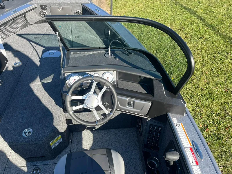 Slide: The Image of 2025 Lund 1875 Pro-V boat cockpit with steering wheel and dashboard, on grassy background. - 15