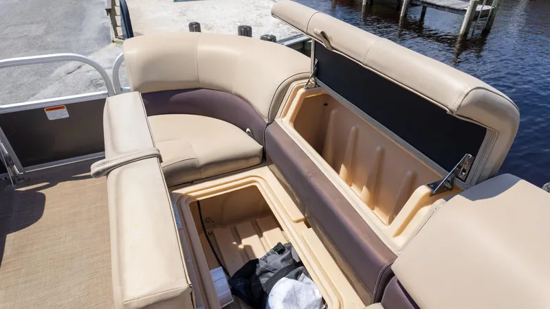 Slide: The Image of 2019 Sun Tracker 24 DLX Party Barge with open storage compartment, beige seating, and waterfront view. - 24