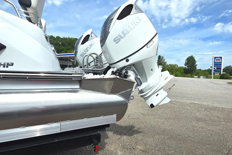 Slide: The Image of 2025 Manitou 27 boat with Suzuki outboard motors, parked outdoors under a blue sky. - 2